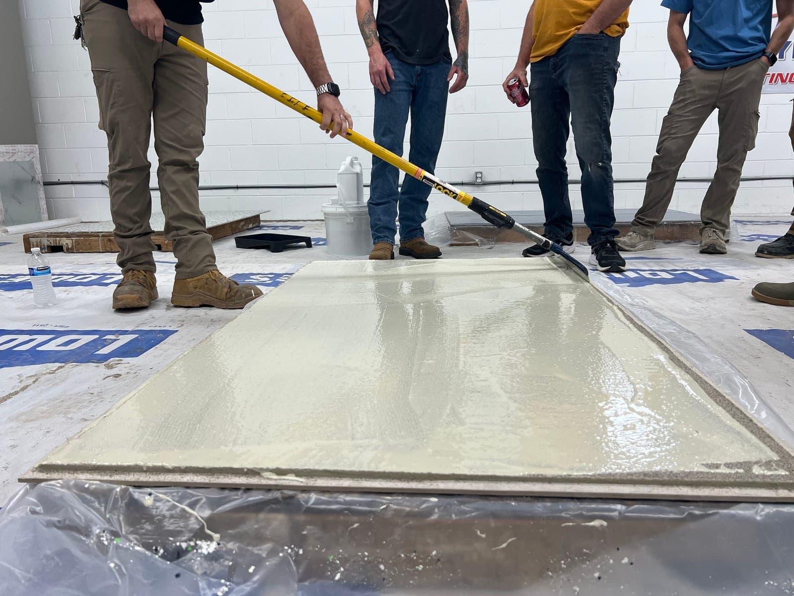 Training group gathered close around a freshly poured panel as one participant pulls a squeegee pass across it